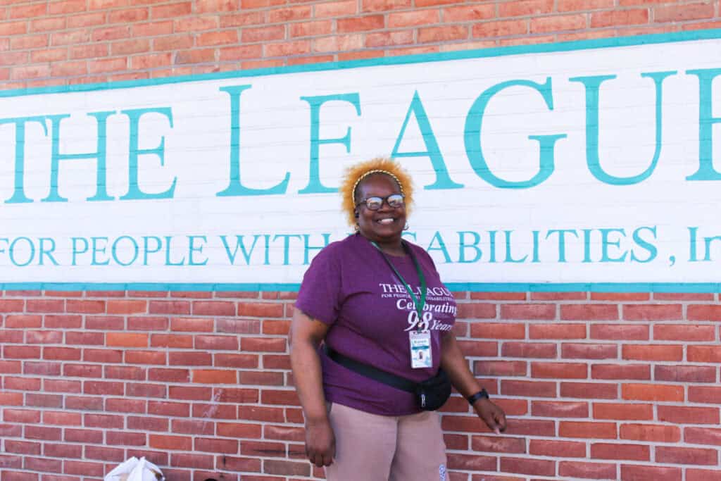 A 64-year old black woman wearing glasses, a maroon t-shirt, and khaki pants stands smiling in front of a brick wall with The League's white and green logo painted on it.