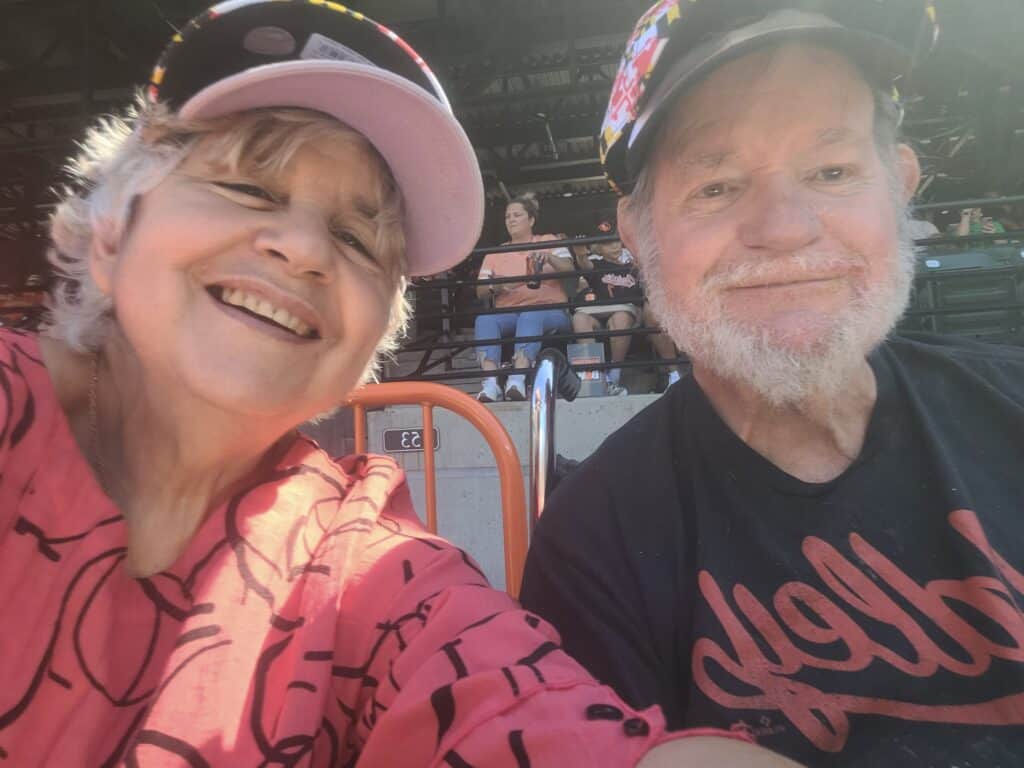 A close up picture of an older white woman wearing a pink baseball cap with her husband at an Orioles baseball game.