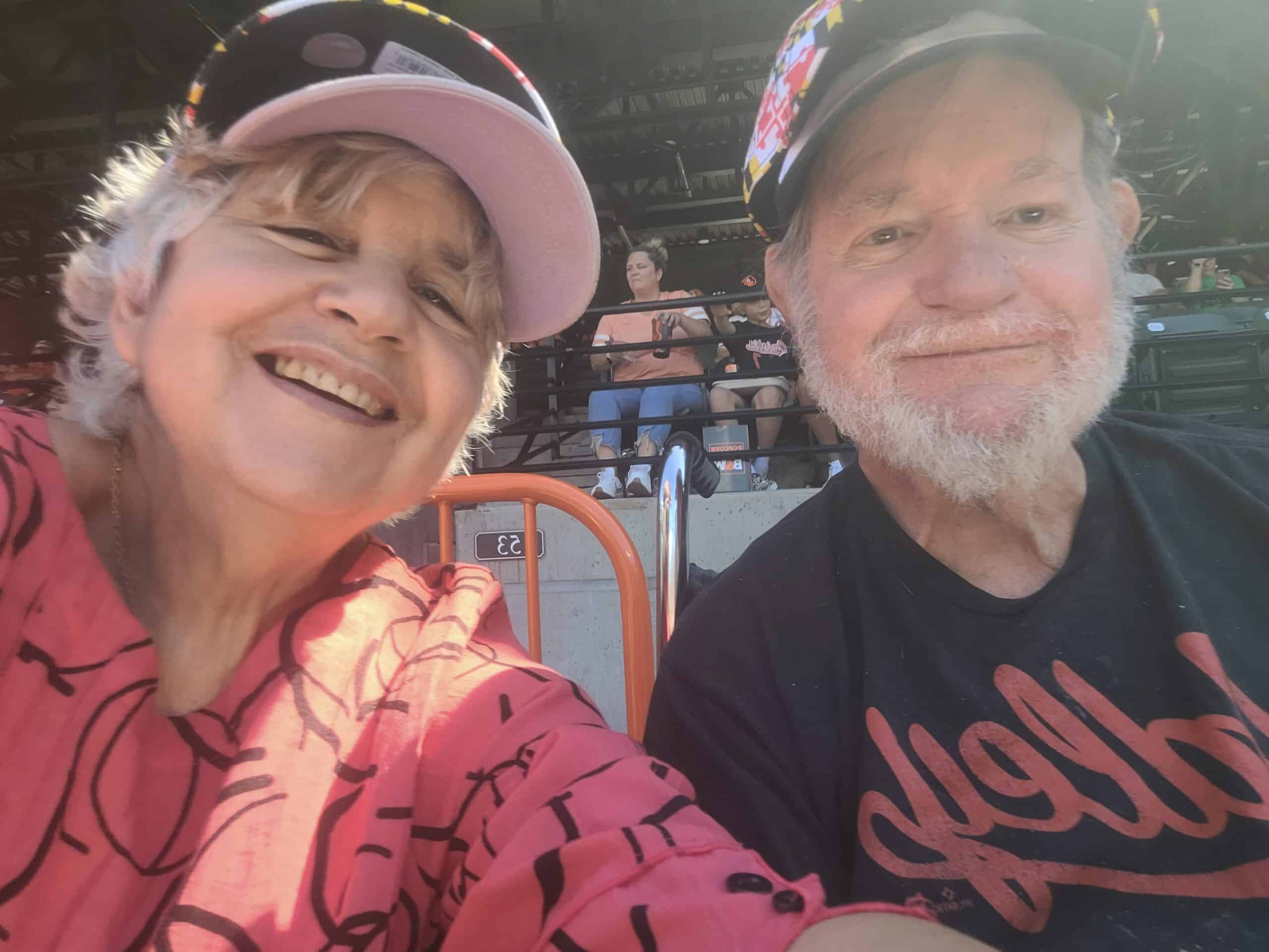 A close up picture of an older white woman wearing a pink baseball cap with her husband at an Orioles baseball game.