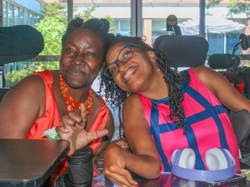 Two black women in power wheelchairs smiling for a photo together at a social event