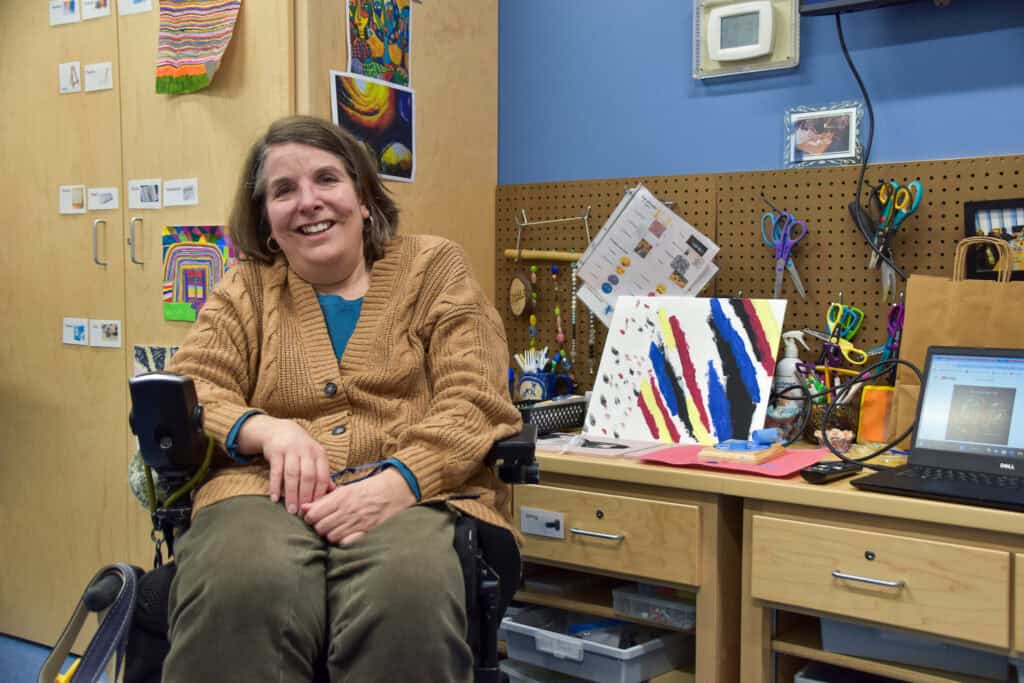 A woman in a tan sweater and blue shirt posing with her art