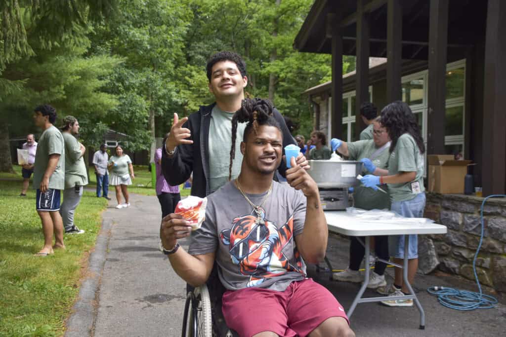 A black man with dreads wearing a gray graphic t-shirt and sitting in a wheelchair holds a blue snowball with a support counselor standing behind him.