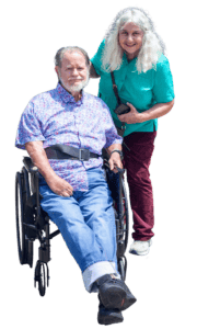 A white man in a short-sleeve button down shirt and blue jeans seated in a wheelchair next to his wife wearing a green t-shirt and brownish-red pants. 