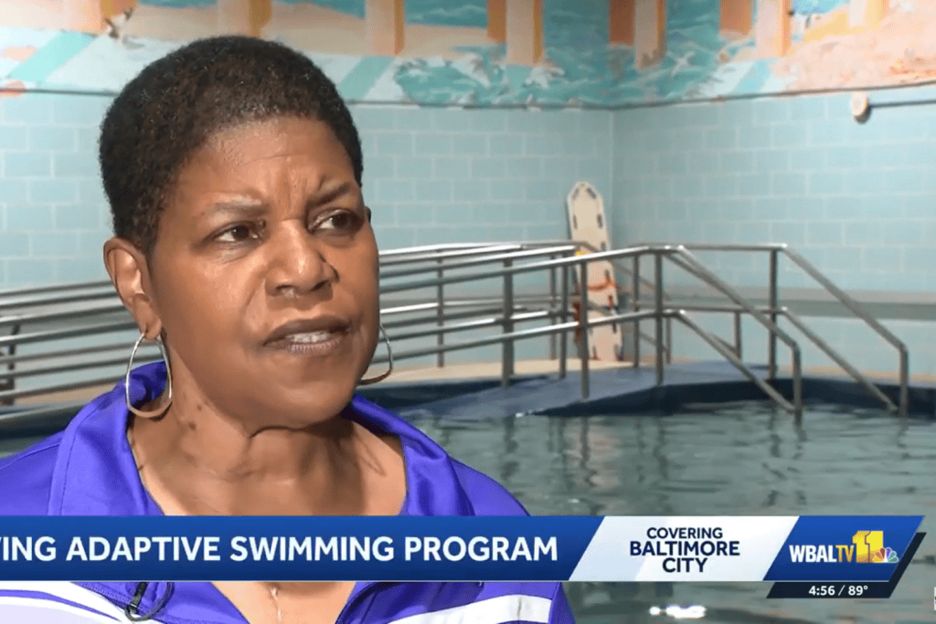 A woman wearing a track suit stands in front of an indoor pool with a seaside mural on the wall for a TV interview.